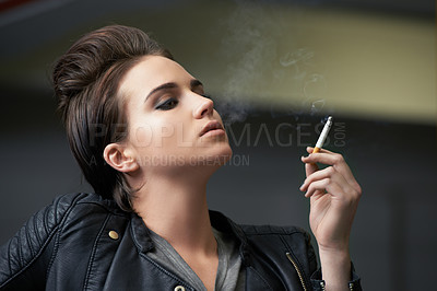 Buy stock photo Shot of a rebellious looking young woman smoking a cigarette in a car garage