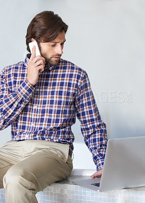 Buy stock photo A young man working on his laptop while talking on the phone