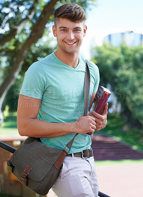 Buy stock photo A handsome student standing outside on campus with a satchel and a textbook