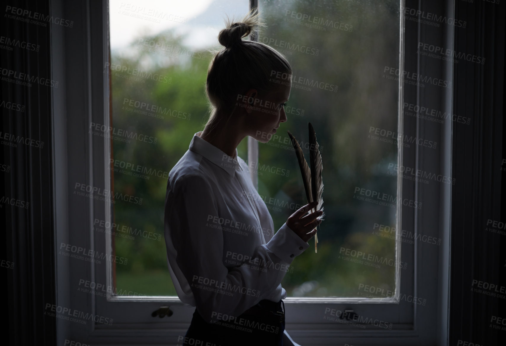 Buy stock photo Woman, feather and window in home for writing in dark room and mysterious aesthetic for artistic pose. Female writer, dim light and ink pen with thinking, author and old school setting for creativity