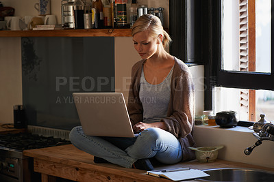 Buy stock photo Shot of a young woman sitting on her kitchen counter using a laptop