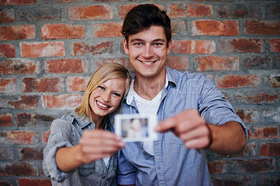 Buy stock photo A young couple standing in front of a brick wall holding a photo to the camera
