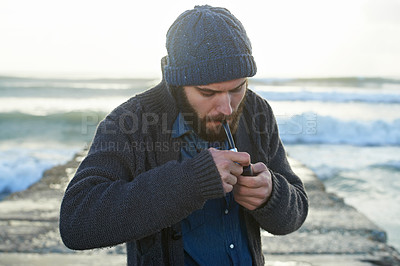 Buy stock photo Bearded, man and smoking a pipe by ocean, lighter and tobacco habit on winter morning for sunrise. English guy, nicotine and vintage smoker for calm, satisfaction and vacation on beach in cape town