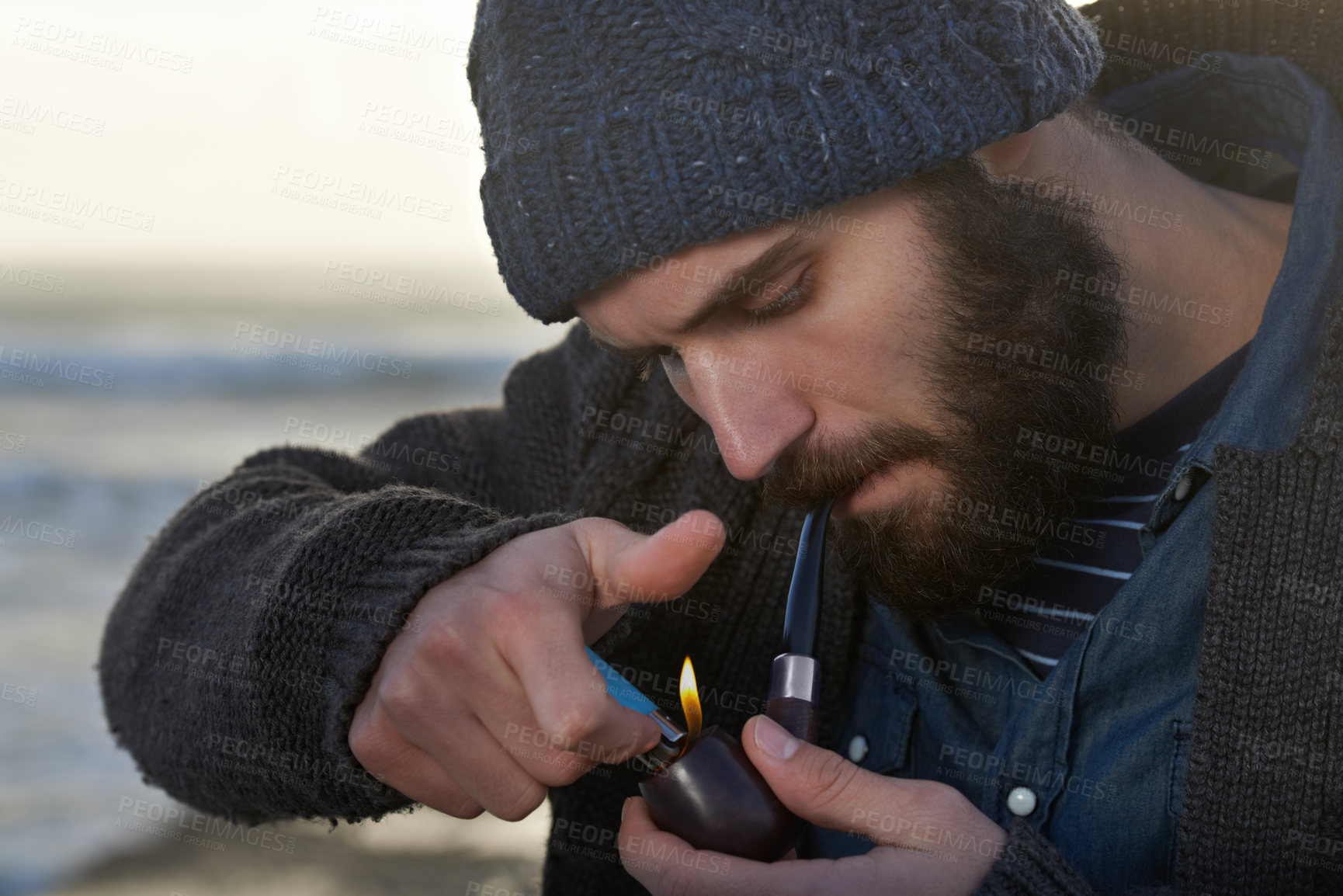 Buy stock photo Bearded, man and smoking a pipe on beach, lighter and tobacco habit on winter morning for sunrise. English guy, nicotine and vintage smoker for calm, satisfaction and vacation by ocean in cape town