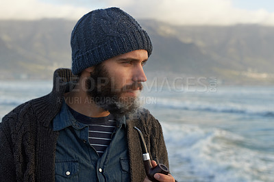 Buy stock photo Beard, man and smoking a pipe by ocean, thinking and sailor habit on winter morning to relax. English person, nicotine and vintage smoker with breathe smoke, calm and vacation in cape town by beach