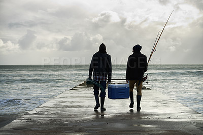 Buy stock photo Shot of two young men fishing at the ocean in the early morning