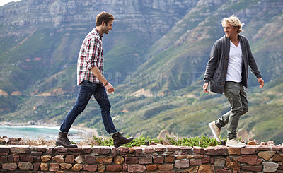 Buy stock photo Shot of two young men walking on a stone wall with the mountain in the background