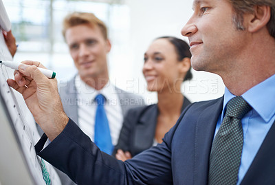 Buy stock photo A businessman writing on a flip-chart as his colleagues stand by