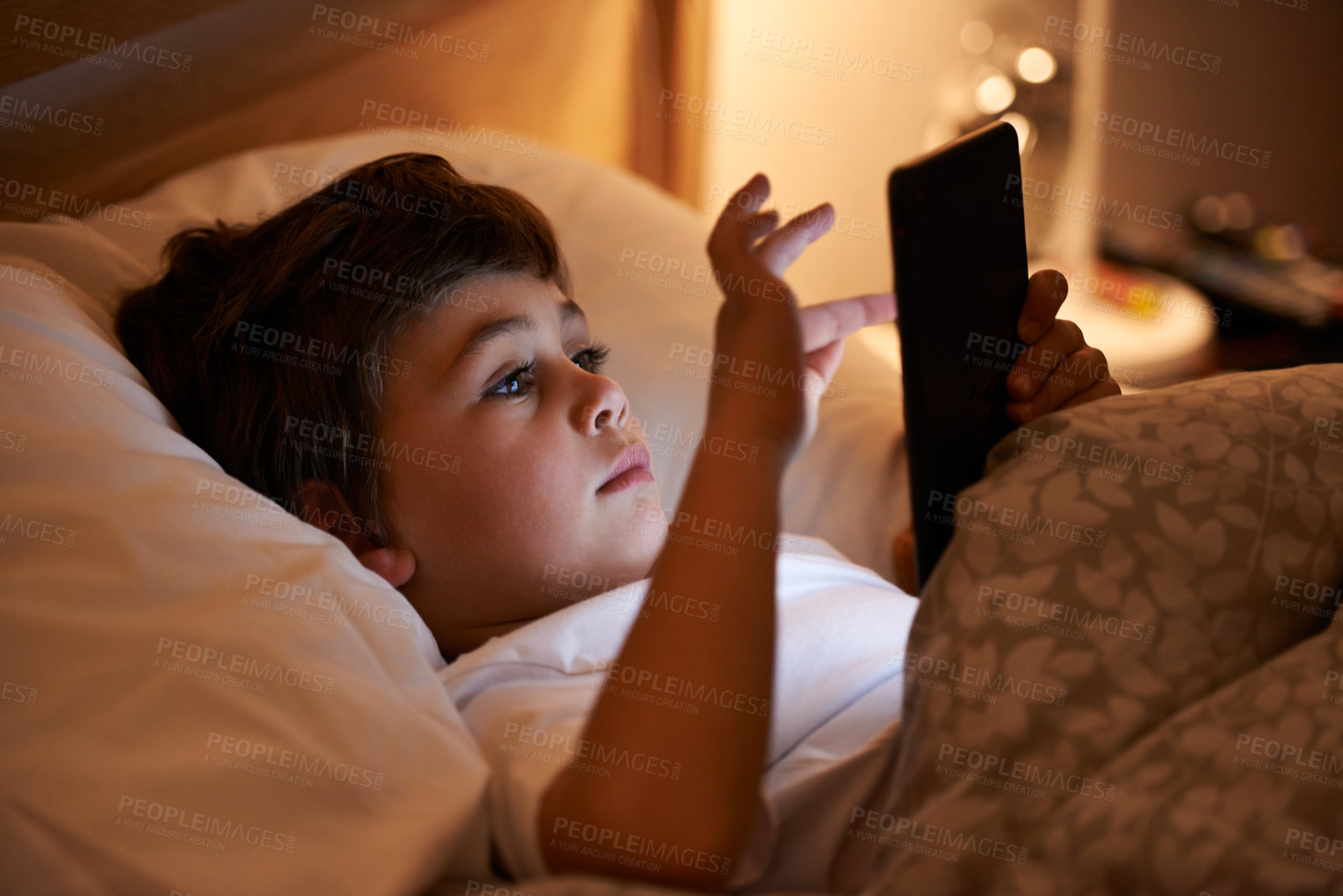Buy stock photo Shot of a young boy playing on a tablet while lying in bed