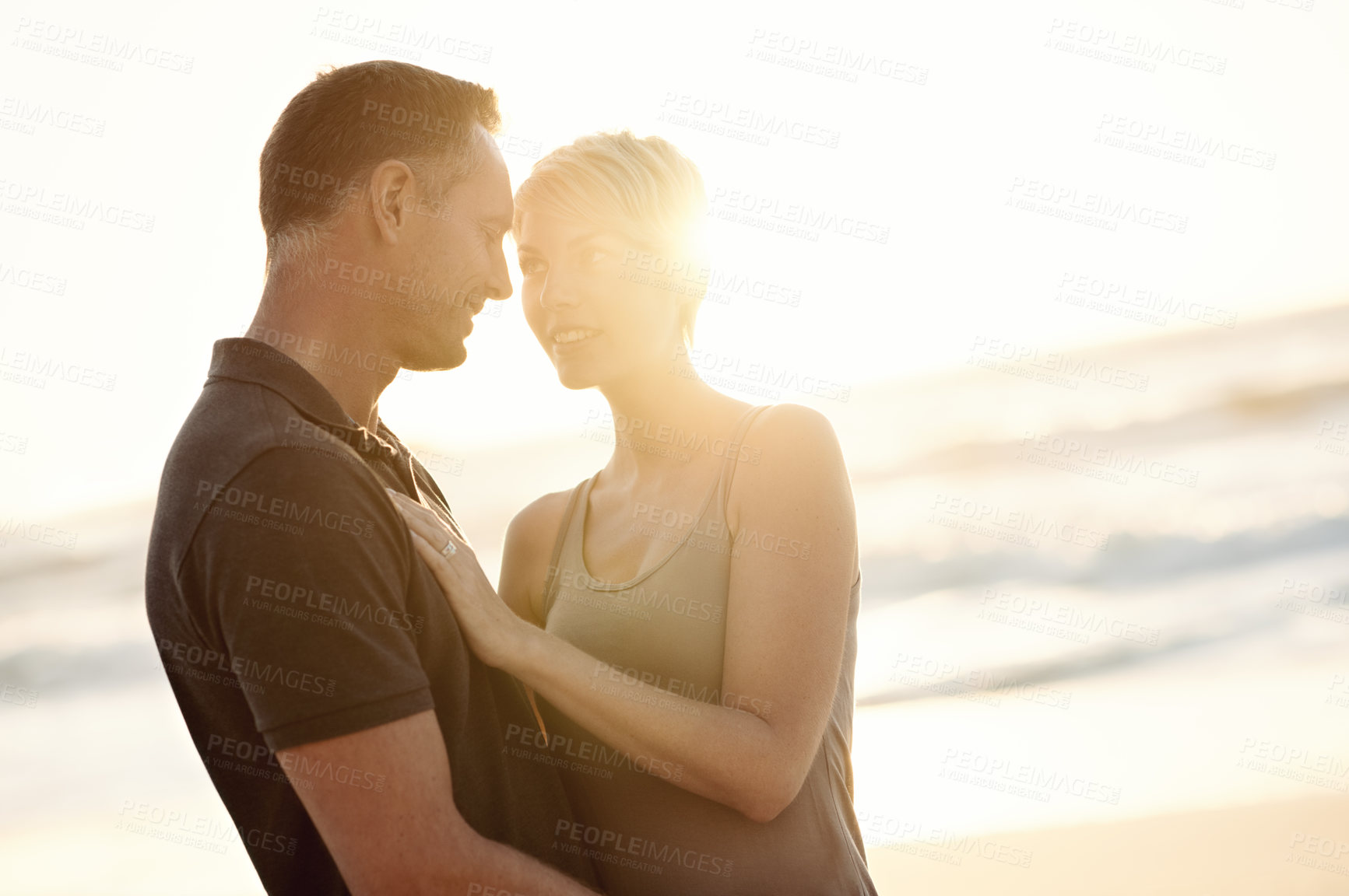 Buy stock photo Couple, face and happy on beach with love for bonding, honeymoon date and weekend holiday with sunset. Lens flare, man and woman with embrace, smile and care for vacation, travel and adventure at sea