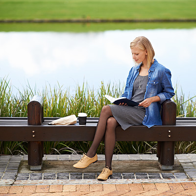 Buy stock photo Shot of an attractive blonde woman reading a book during her lunch break on a park bench
