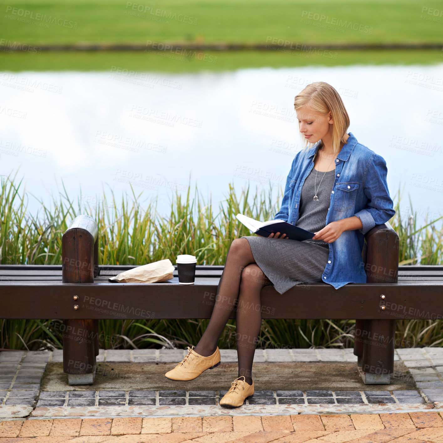 Buy stock photo Shot of an attractive blonde woman reading a book during her lunch break on a park bench