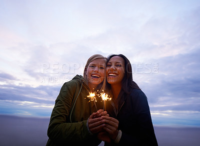 Buy stock photo Two young women holding sparklers with the ocean and twilight sky behind them