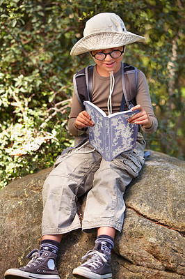 Buy stock photo Happy, little boy and reading in nature with book for travel guide, adventure or outdoor discovery. Male person, child or kid with glasses, hat and notebook on rock for story or exploration in forest