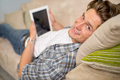 Buy stock photo Shot of a handsome young man using his digital tablet while relaxing on the sofa