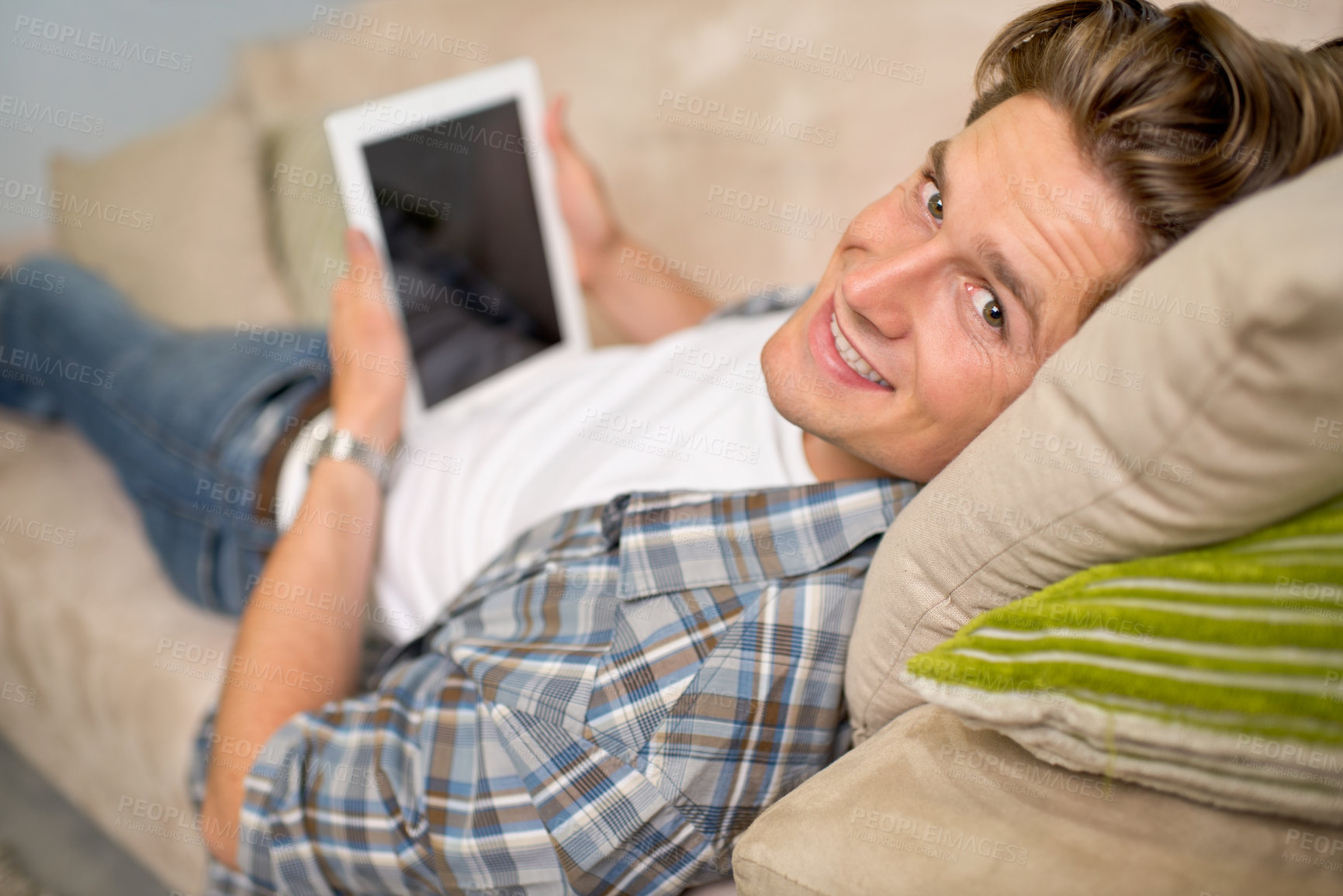 Buy stock photo Shot of a handsome young man using his digital tablet while relaxing on the sofa