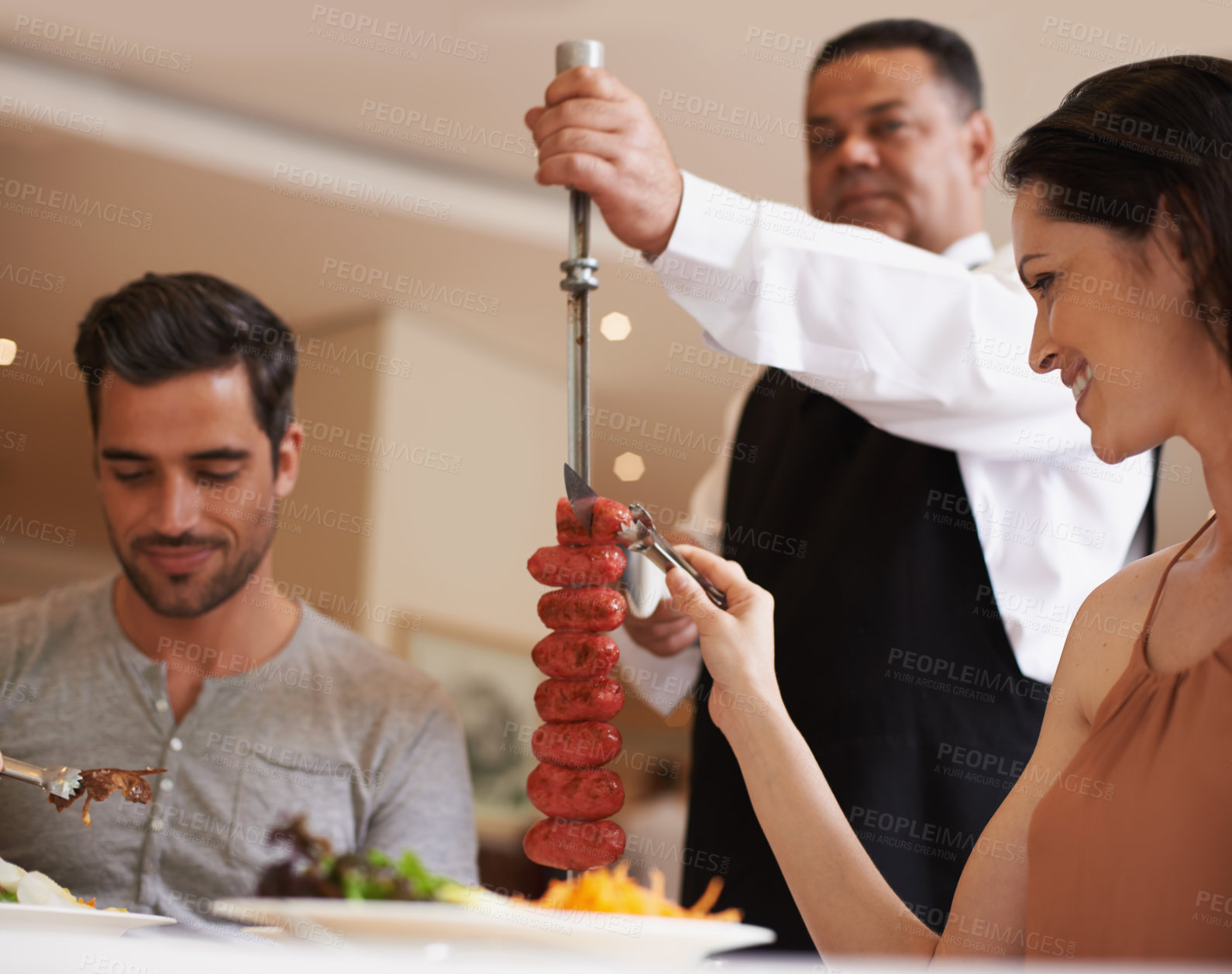 Buy stock photo Couple, waiter and service for lunch in restaurant with meat, happiness and fine dining for anniversary or honeymoon. Man, woman and employee with sausage on skewer, healthy meal and table in London