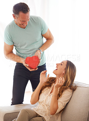 Buy stock photo Husband, surprise and giving gift to wife in home, excited and marriage to celebrate valentines day on couch. Man, love and care for woman with kindness, gratitude and romantic with a heart present