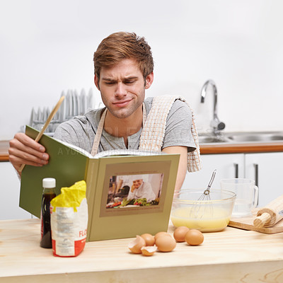 Buy stock photo A young man standing in his kitchen and looking perplexed while reading a recipe book 