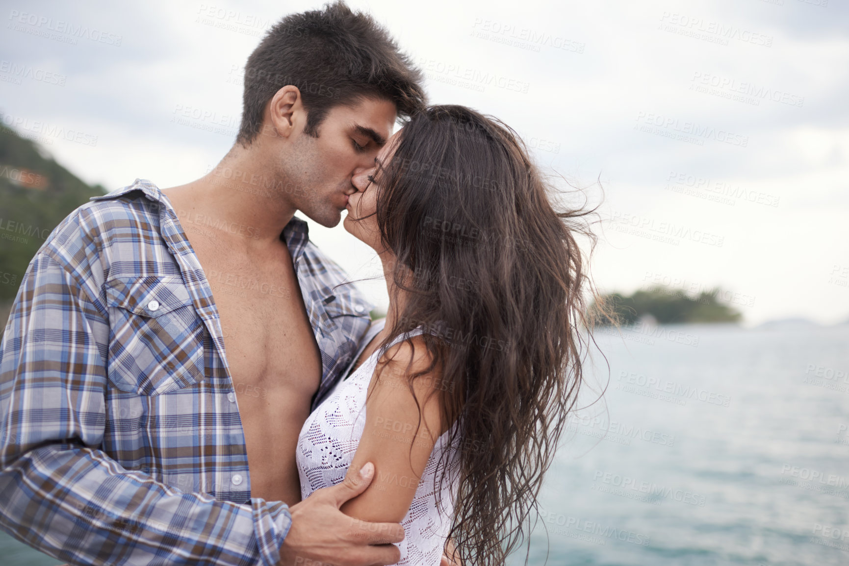 Buy stock photo Shot of an intimate young couple enjoying a kiss by the water's edge