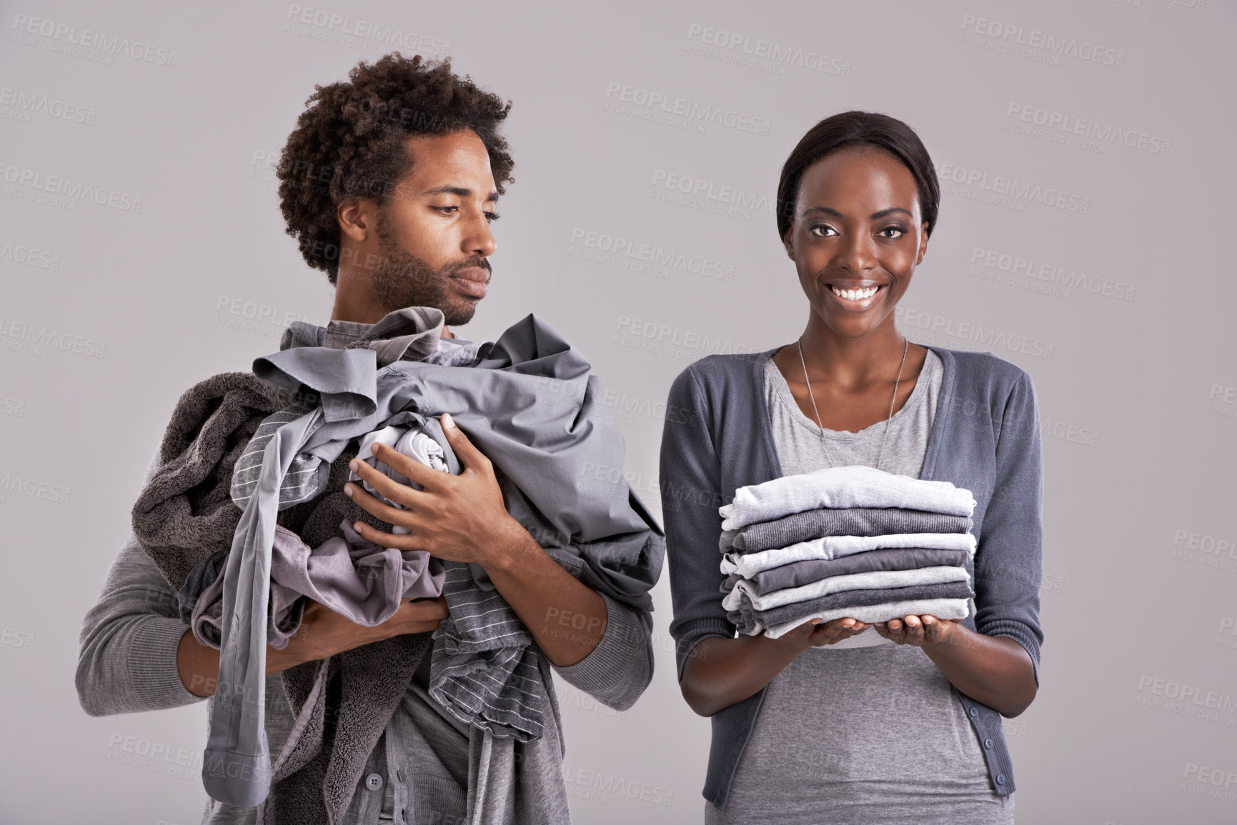 Buy stock photo People, studio and pile of clothes in hand for laundry day, wash and couple cleaning together. Woman, happy and  neat washing folded with messy, man and unhappy for dirty wardrobe on gray background