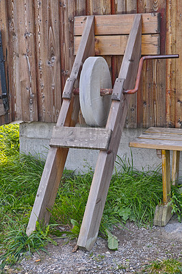 Buy stock photo An antique grindstone standing against a wooden shed with overgrown grass. Old grey sharpening stone tool or equipment used outside on a farm