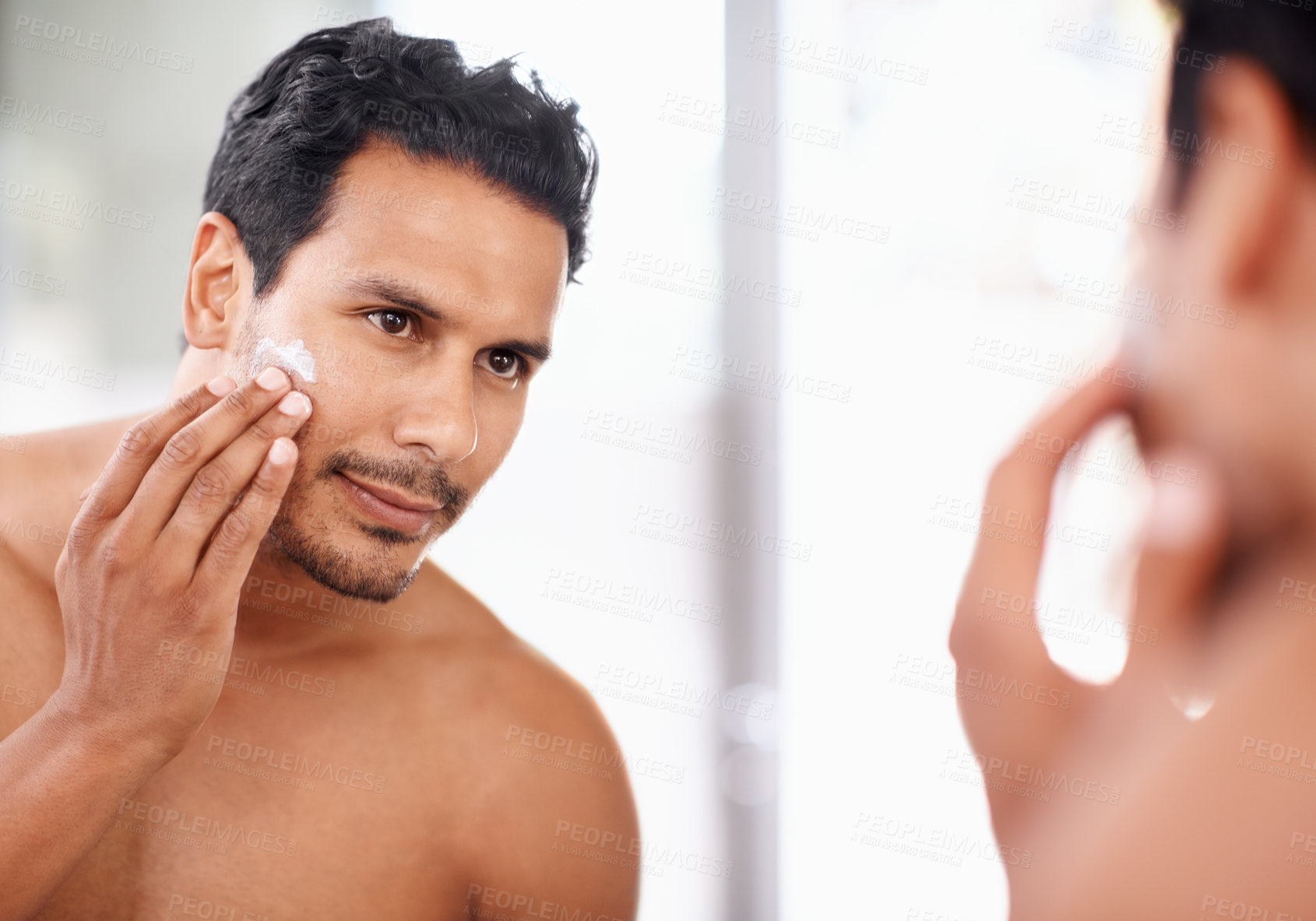 Buy stock photo A young man applying cream to his face while looking in the mirror