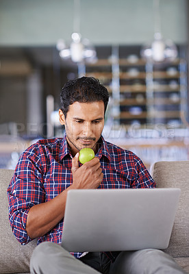 Buy stock photo A young man eating an apple while working on his laptop at home