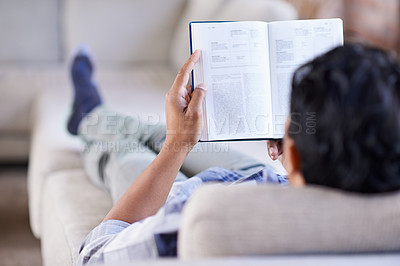 Buy stock photo Rearview shot of a young man reading a novel while relaxing on the couch at home