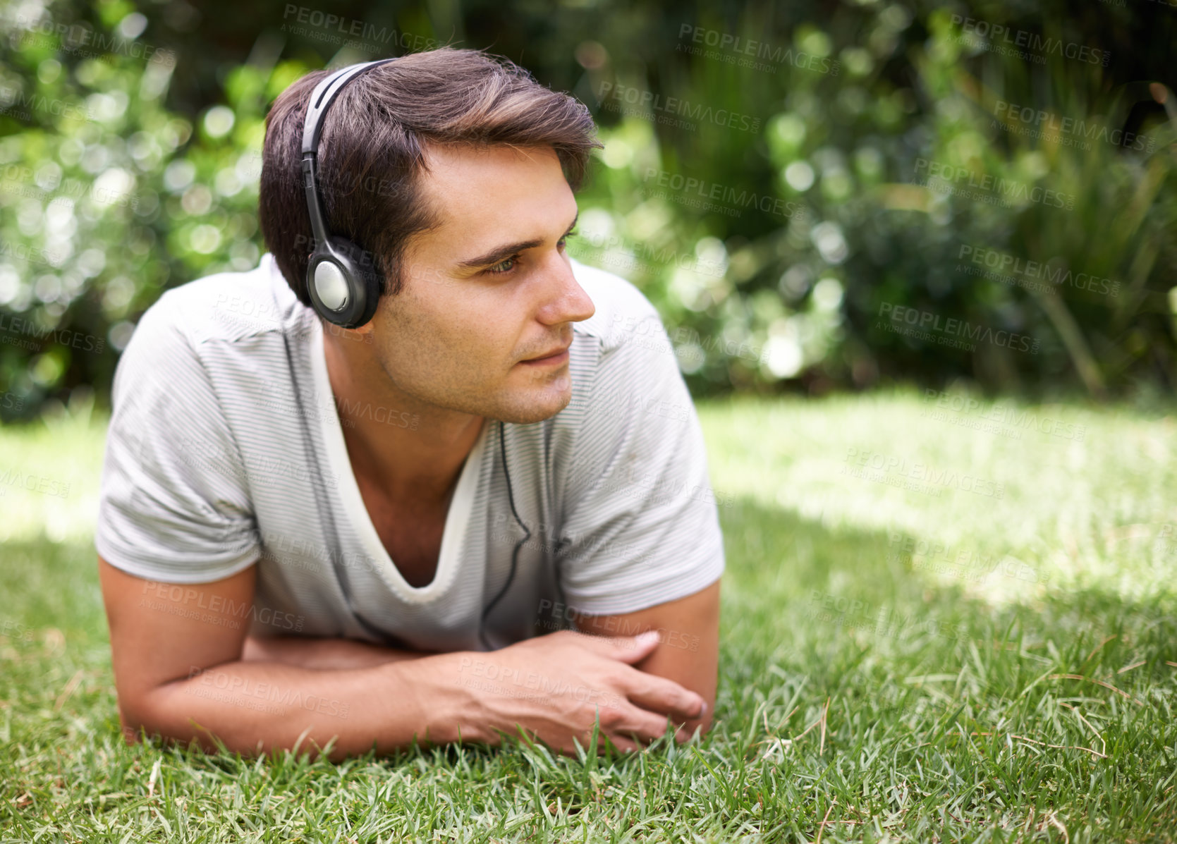 Buy stock photo Handsome young man wearing headphones and listening to music with while lying on the green grass at a park in summer. Carefree male looking content while relaxing outdoors and enjoying his free time