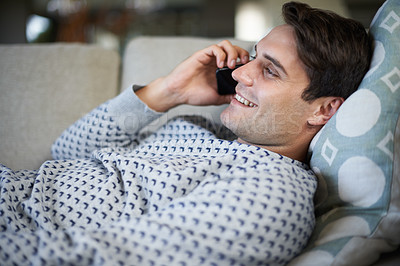 Buy stock photo Shot of a young man lying on his sofa while talking on his phone
