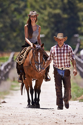 Buy stock photo An attractive young cowgirl riding a horse on a ranch while a cowboy walks alongside