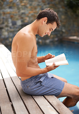 Buy stock photo Cropped shot of a young man reading a book while sitting by the pool