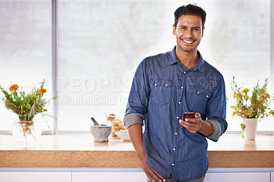 Buy stock photo Portrait of a handsome holding a mobile phone while standing in the kitchen