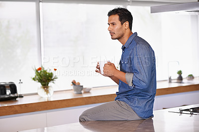 Buy stock photo A serious man holding a mug of coffee and sitting on his kitchen counter