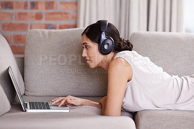 Buy stock photo Woman, laptop and lying on sofa with headphones for streaming, music or search for web video in home. Girl, computer or scrolling for movie, podcast or audio on internet to relax on living room couch