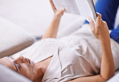 Buy stock photo Shot of a young woman reading a novel while lying on a sofa