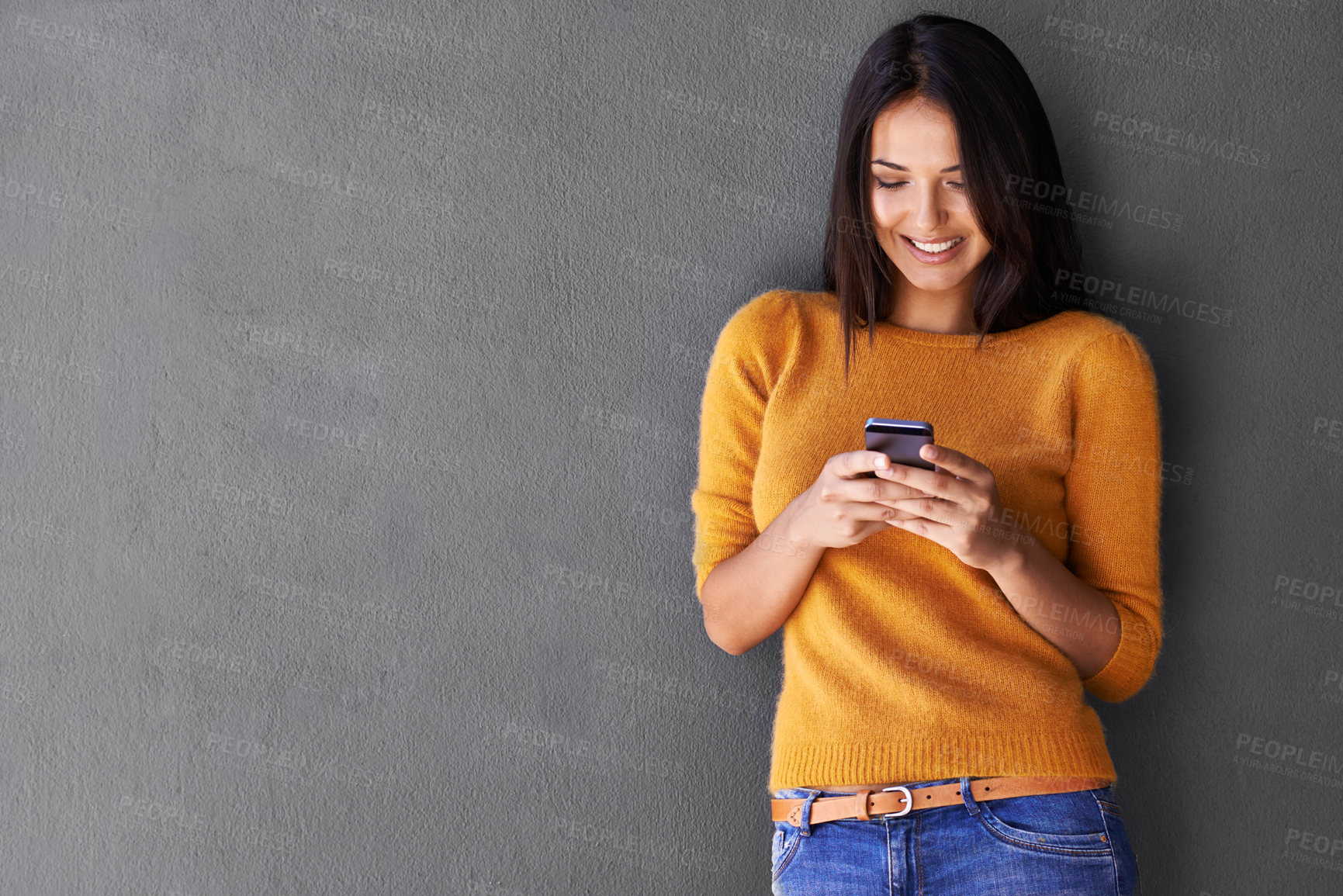 Buy stock photo An attractive young woman using a mobile phone while standing against a gray wall