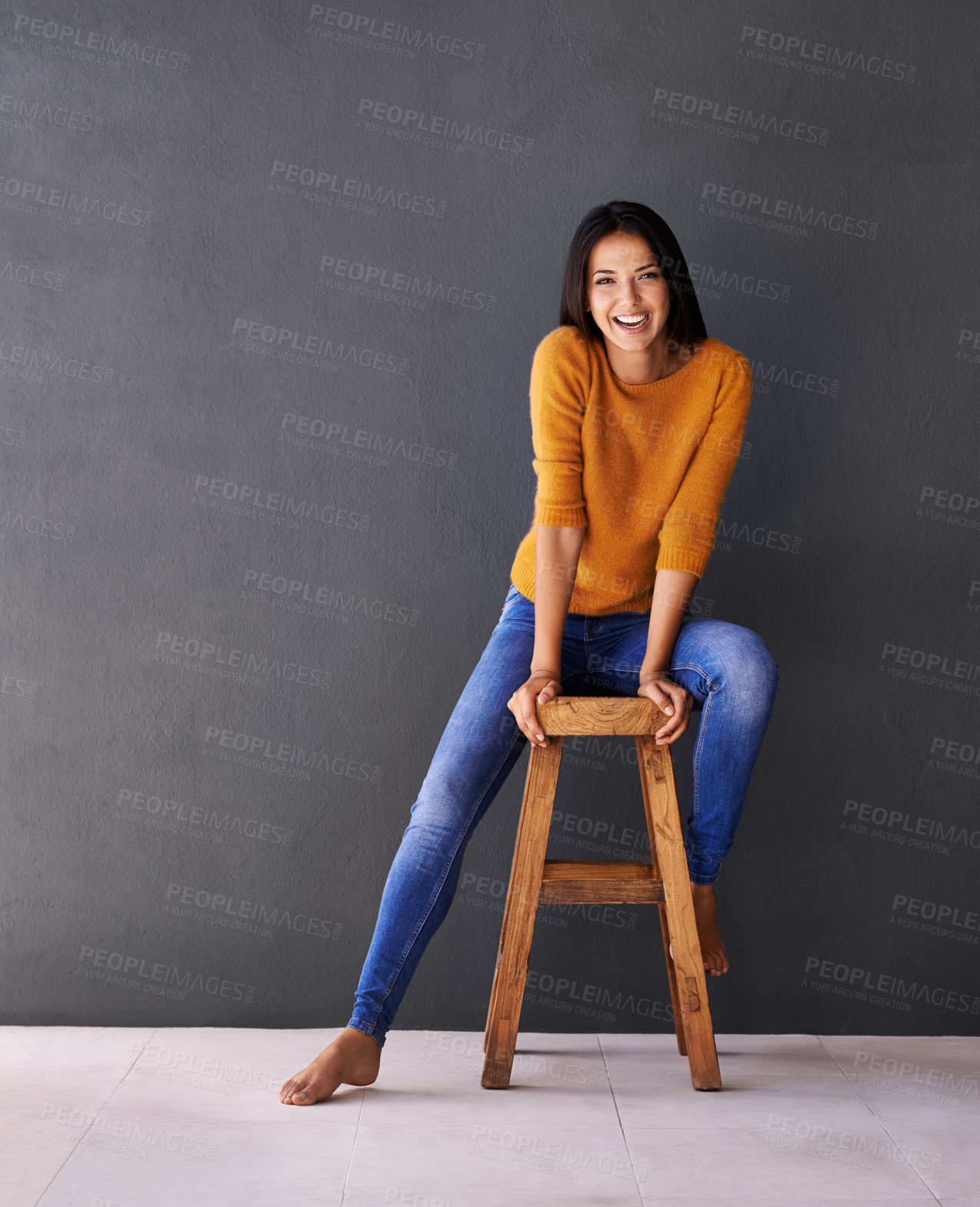 Buy stock photo Portrait of a happy young woman sitting on a stool against a gray wall