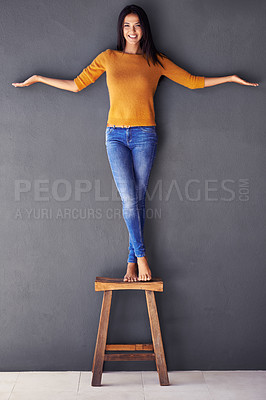 Buy stock photo Woman, fashion and stand on chair in portrait, smile and balance on wall background. Female person, modern clothing and happy lady on stool, barefoot and confidence in living room at home in house