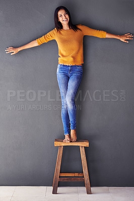 Buy stock photo Woman, confident and smile on chair in portrait, stand and fashion on wall background. Female person, studio and happy lady on stool, barefoot and balance in living room at home with modern clothing