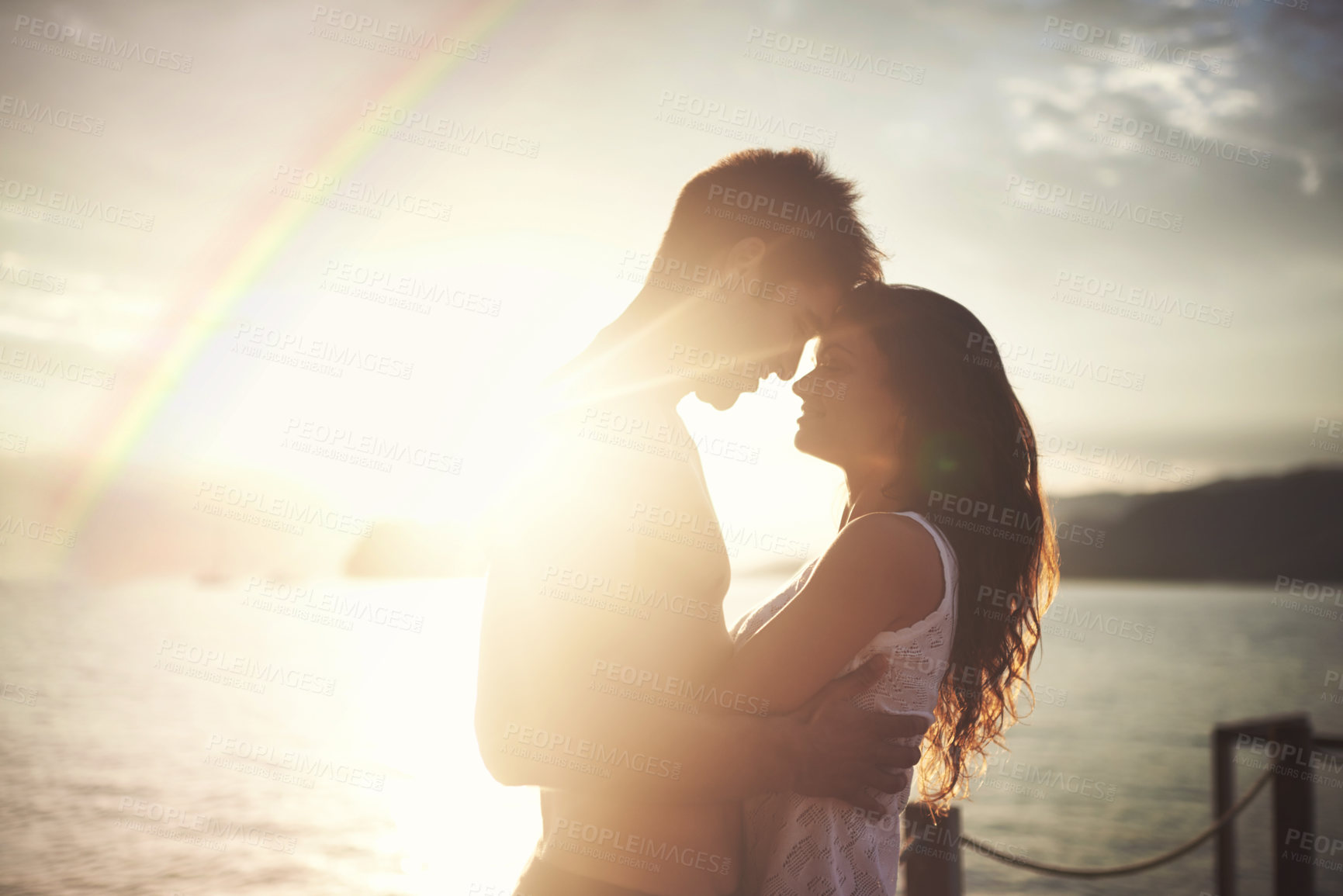 Buy stock photo Couple, ocean and holding each other at sunset, loving and smile on honeymoon. Rainbow, water and shoreline during holiday celebration, married and romantic getaway with sunshine flare in horizon 

