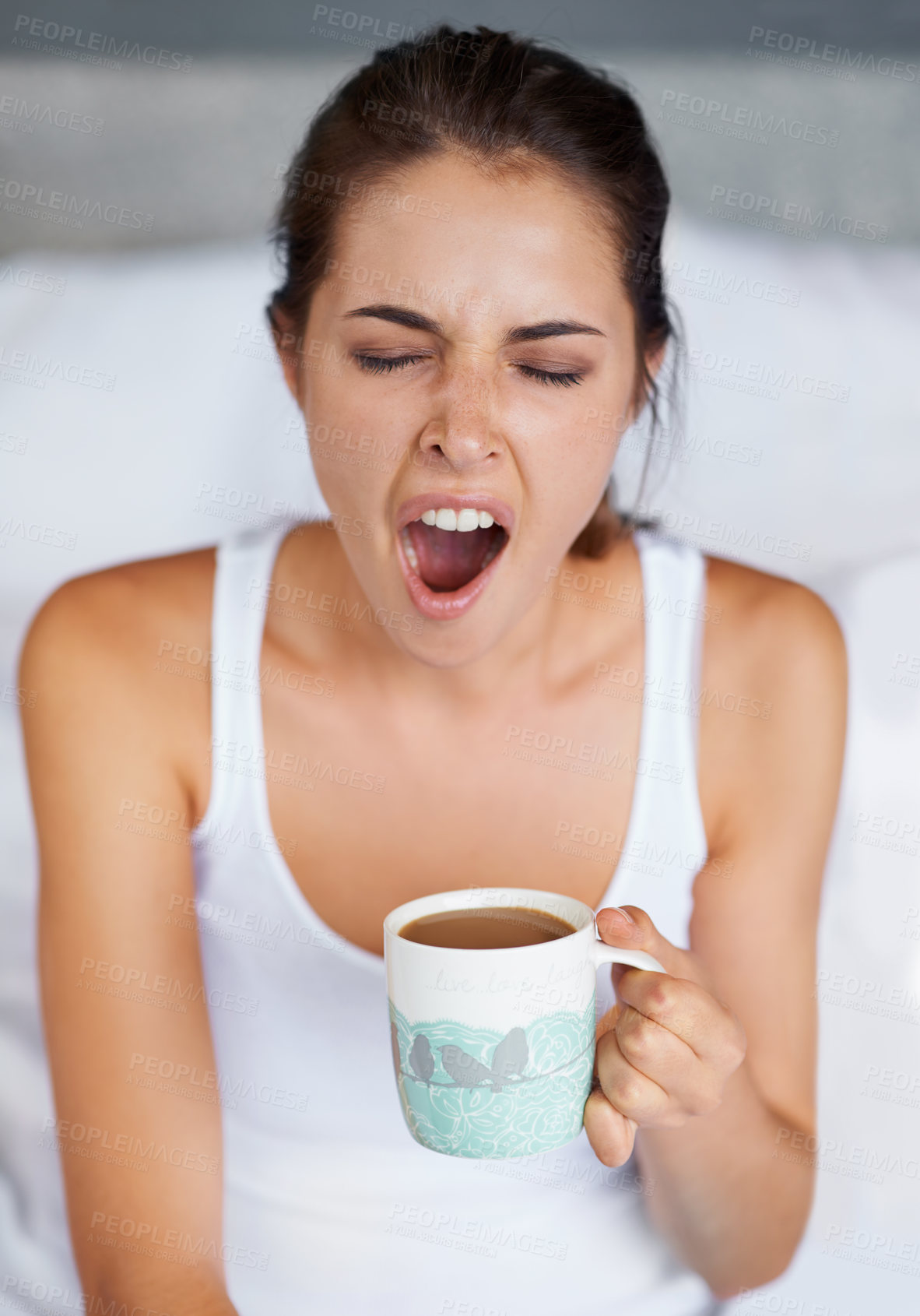 Buy stock photo A young woman giving a big yawn while sitting with a cup of coffee in bed