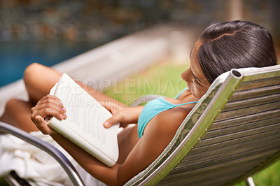 Buy stock photo A young woman reading a book while relaxing in a lounge chair by a swimming pool