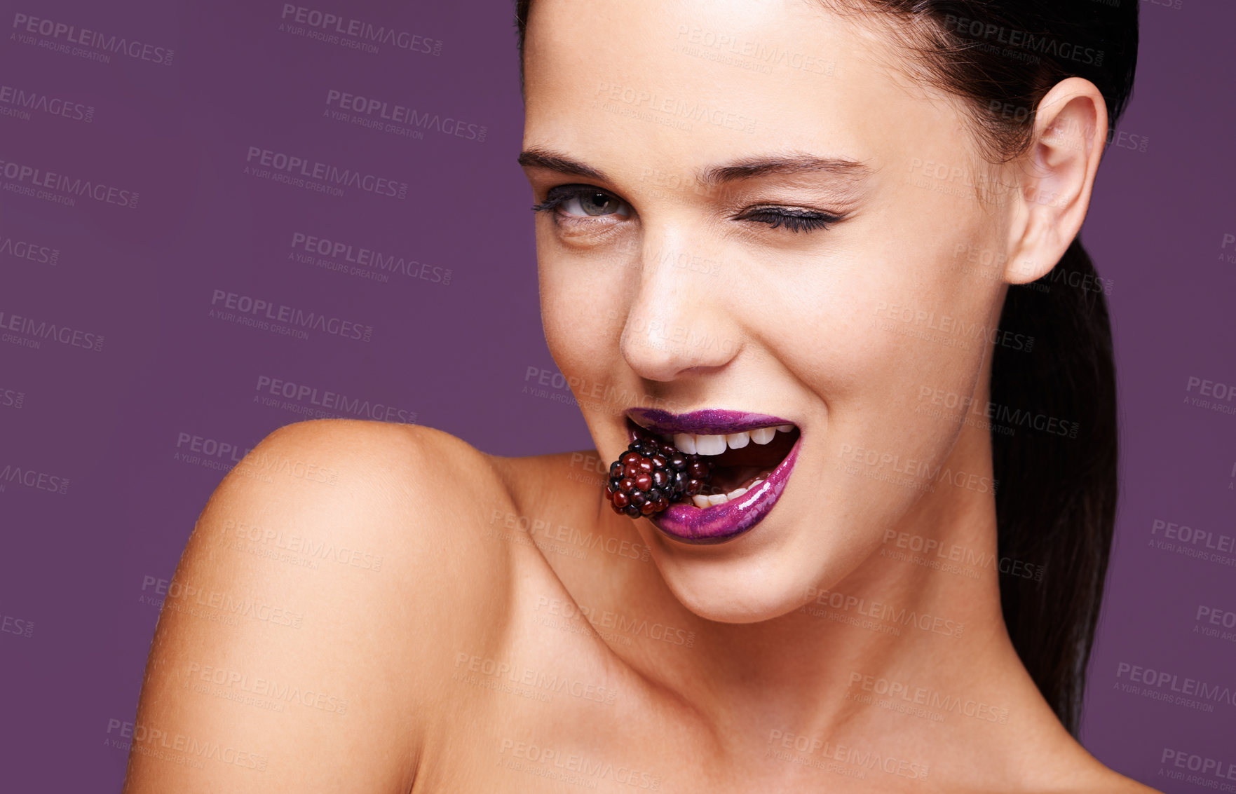 Buy stock photo A studio portrait of a beautiful young woman wearing purple lipstick and biting into a berry