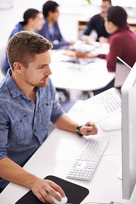 Buy stock photo Shot of a young designer working at his computer with colleagues in the background