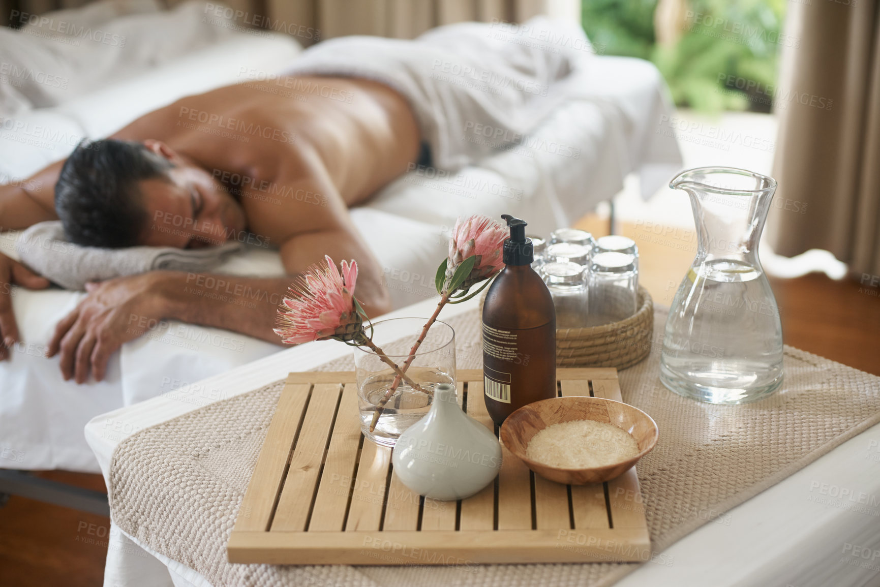 Buy stock photo Salt, flowers and man in spa to relax on bed or break with luxury pamper treatment tools on table. Protea, facial oil or person at resort and salon for natural healing benefits or massage with water