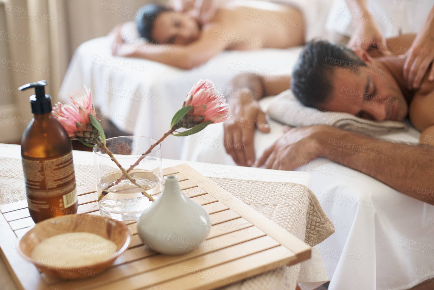 Buy stock photo Hotel, flowers and couple in spa to relax on bed or break with luxury pamper treatment tools on table. Protea, facial oil or woman with man at resort or salon for natural healing benefits or massage