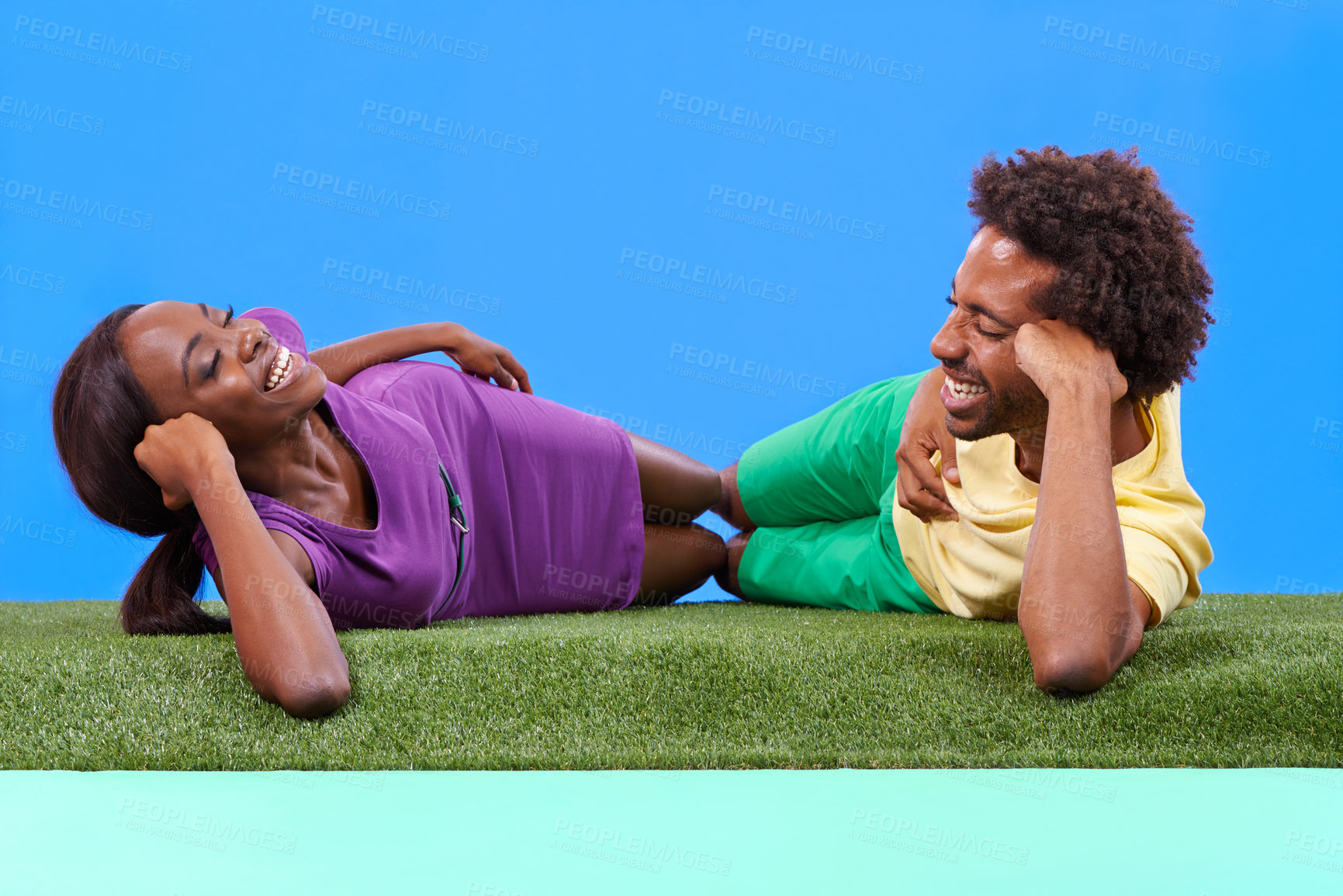 Buy stock photo Happy, black couple and lying with green grass for love, romance or outdoor weekend against a blue studio background. African man and woman laughing in relax for fun holiday bonding on mockup space