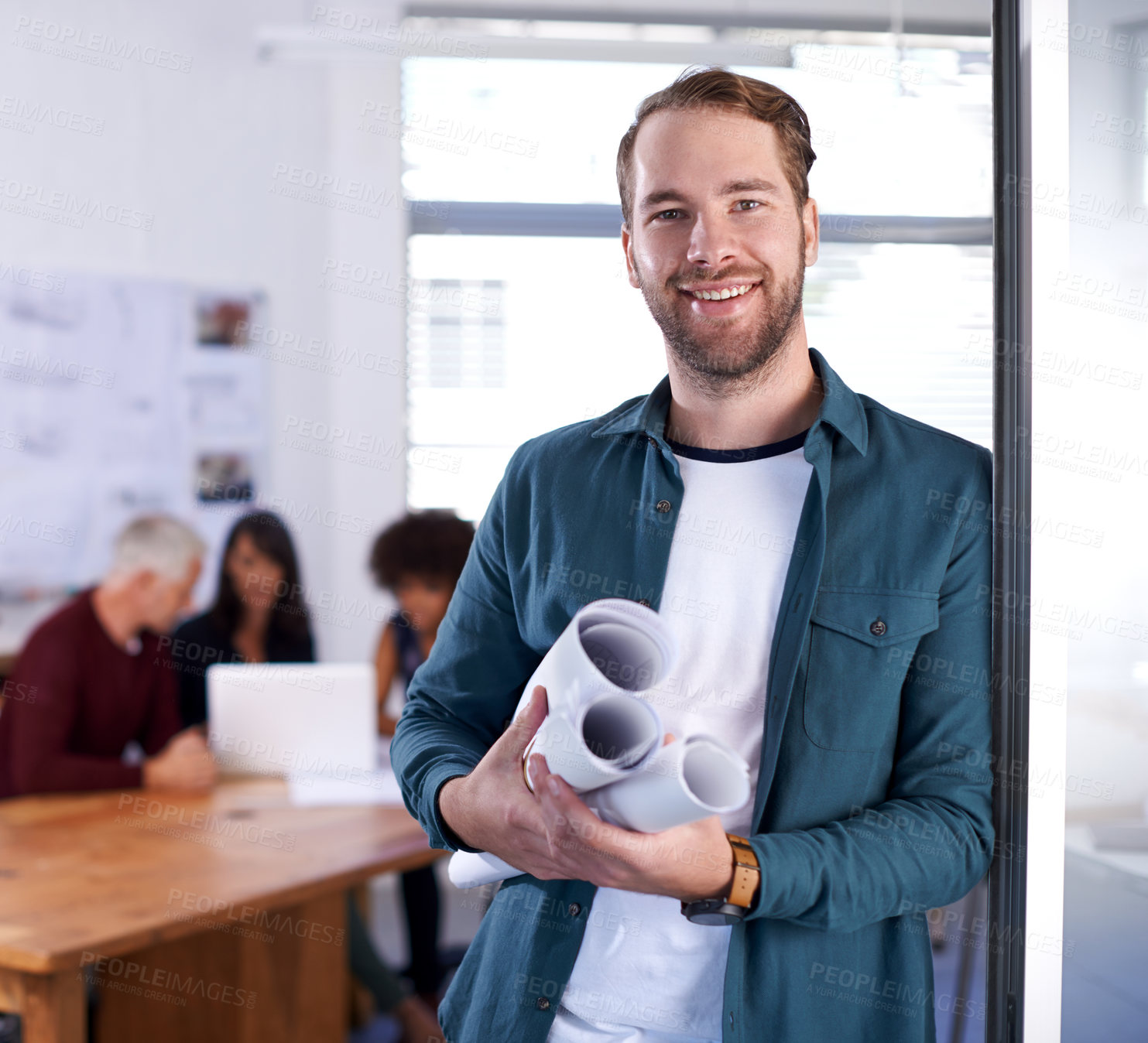 Buy stock photo Portrait of a young architect holding blueprints with his colleagues working in the background